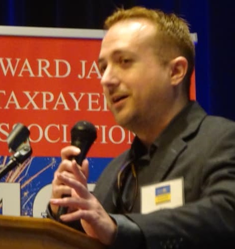 Eric Eisenhammer speaking and gesturing at lectern with microphone portrait - with banner in background with text 'howard jarvis taxpayers association' - wearing dark grey blazer (color photo)